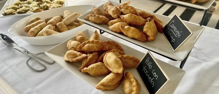 Catering trays of empanadas labeled ground beef, shrimp and chicken with bacon, beside a bowl of tequeños