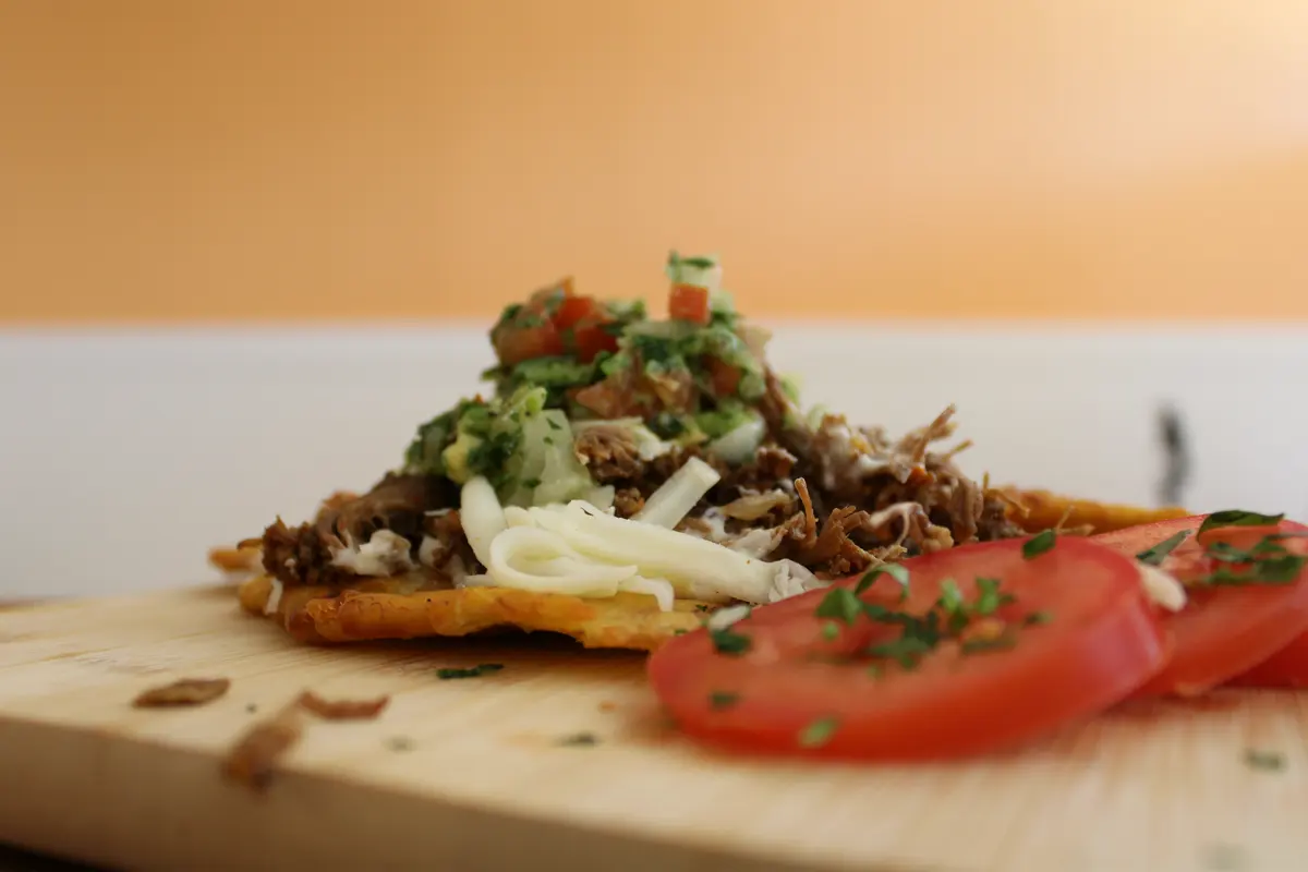 A patacón topped with shredded beef, cheese and pico de gallo, with sliced tomato